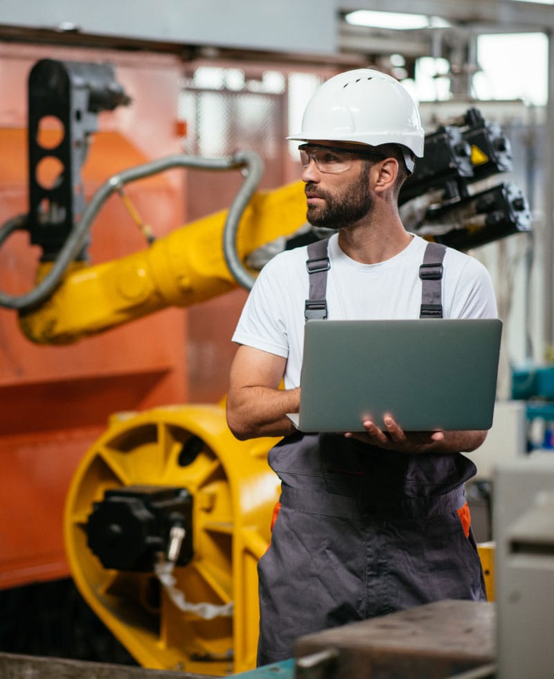 Worker in a factory using a laptop near robotic machinery