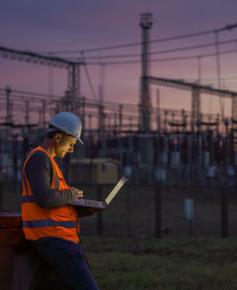 Utility engineer in hard hat and safety vest monitors a laptop at an electrical substation at dusk.